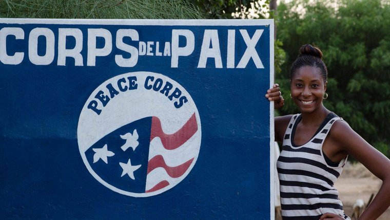 Black woman Peace Corps Volunteer smiles next to Peace Corps sign in French.