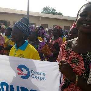 A group of women stand, holding a banner with the Peace Corps logo. One woman smiles at the camera.