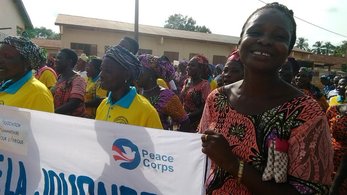 A group of women stand, holding a banner with the Peace Corps logo. One woman smiles at the camera.