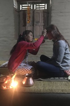 A Volunteer receives a blessing from a Nepali host sister, both are sitting on a floor in a dark room
