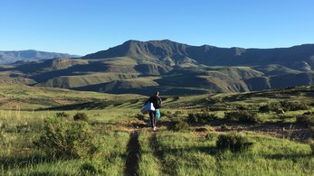 A person walks in the distance through a field in Lesotho. There are mountains in the background and blue skies.