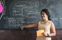 A Puerto Rican woman stands teaching at a blackboard, smiling.