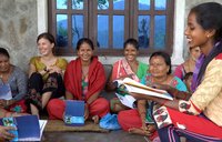 Several Nepalese women and one American volunteer, all in colorful clothing, sit together on a porch reading and smiling.