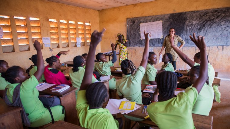 A teacher and a Peace Corps Volunteer stand at the head of a classroom, while students in the class raise their hands to be c