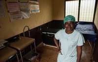 A young female health worker dressed in scrubs stands in exam room.