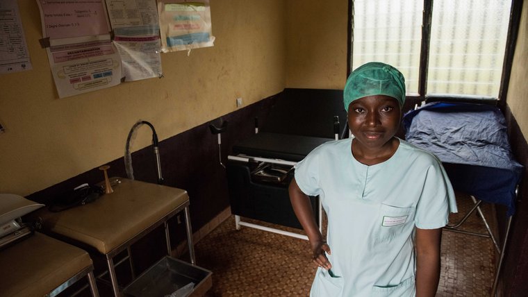 A young female health worker dressed in scrubs stands in exam room.