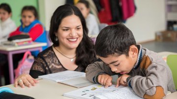 A smiling female Peace Corps Volunteer kneels next to the desk of a young boy reading from a book.