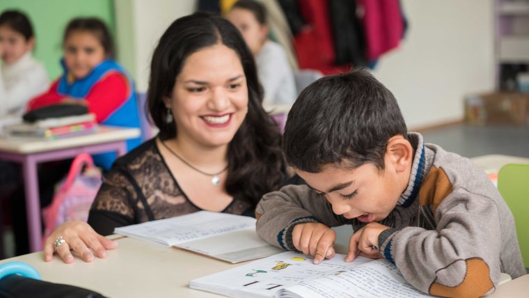 A smiling female Peace Corps Volunteer kneels next to the desk of a young boy reading from a book.