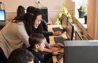 A smiling female Peace Corps Volunteer helps a young boy seated at a computer.
