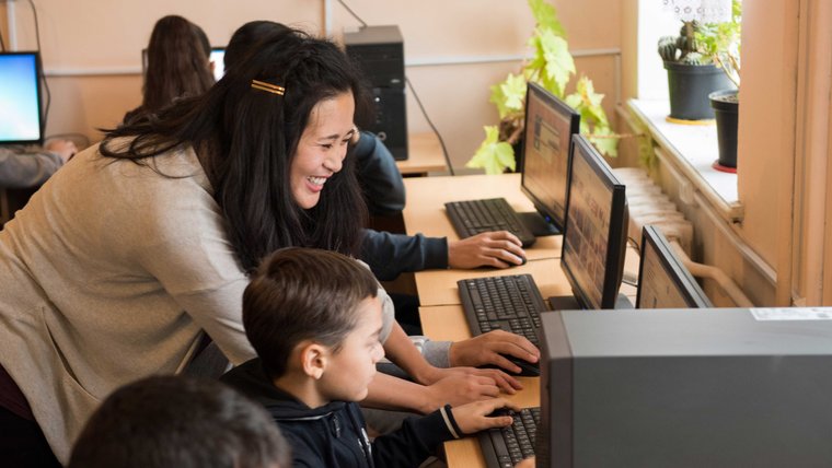A smiling female Peace Corps Volunteer helps a young boy seated at a computer.