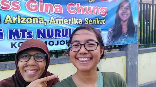 An Asian-American female Volunteer stands next to her Indonesian host mom and smiles in front of a banner with her face on it