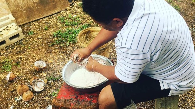 A man sits on a stool making coconut milk in a large metal bowl on the ground. His back is to the camera.