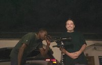 A while male person stand while a black male person is bending, looking through telescope, in the night.