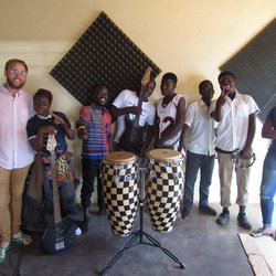 A male Volunteer stands with several Malawian youth, infront of drums and a guitar.