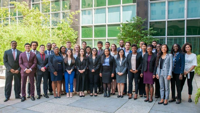 A group of young people in professional clothing stand outside of a building in a large group.
