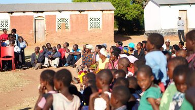 Women, men, and children gather together outside of a school house.