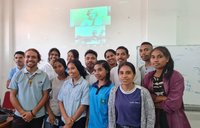 A class of young adults stand in front of a woman displayed through a projector