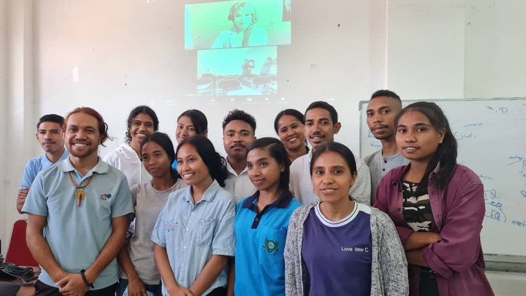A class of young adults stand in front of a woman displayed through a projector