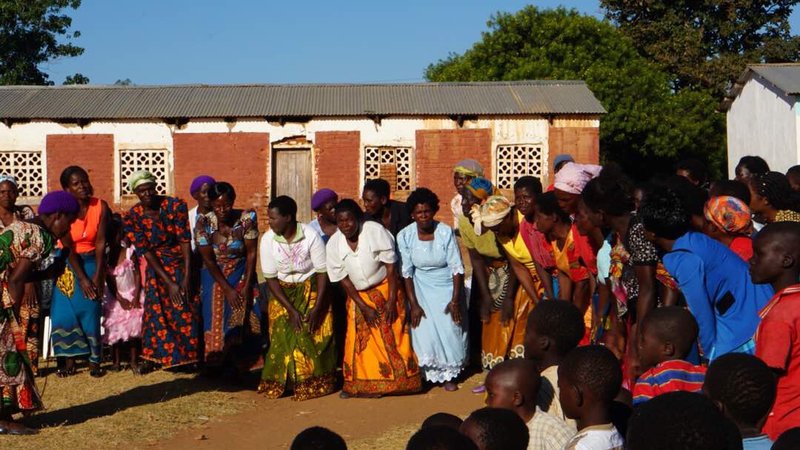 Malawian women with colorful skirts gather to welcome new Trainees outside of a school house