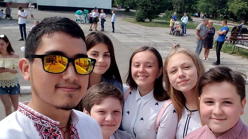 A young Mexican American man wearing traditional Ukrainian clothes stands smiling with his young, Ukrainian students outside.