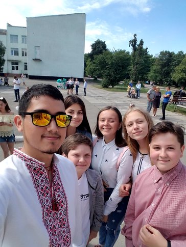 A young Mexican American man wearing traditional Ukrainian clothes stands smiling with his young, Ukrainian students outside.