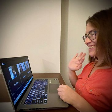 A female in a red shirt waves at the virtual meeting's attendees.