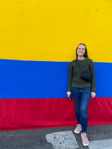 A woman in jeans smiles in front of the yellow, blue, and red Colombian flag.