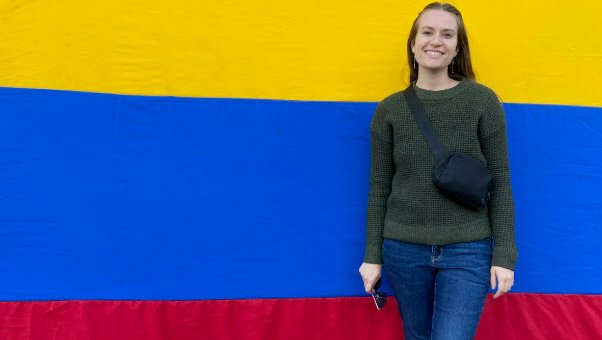 A woman in jeans smiles in front of the yellow, blue, and red Colombian flag.
