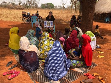 A woman sits holding a visual board while speaking to a group of women seated in front of her.