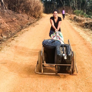 A woman walking along a dirt road, pushing a cart loaded with black containers