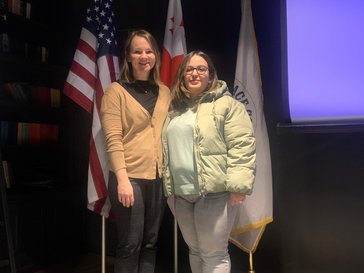 Two women stand in front of three flags.