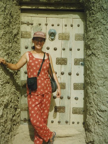 A smiling woman stands in a printed jumpsuit and baseball cap