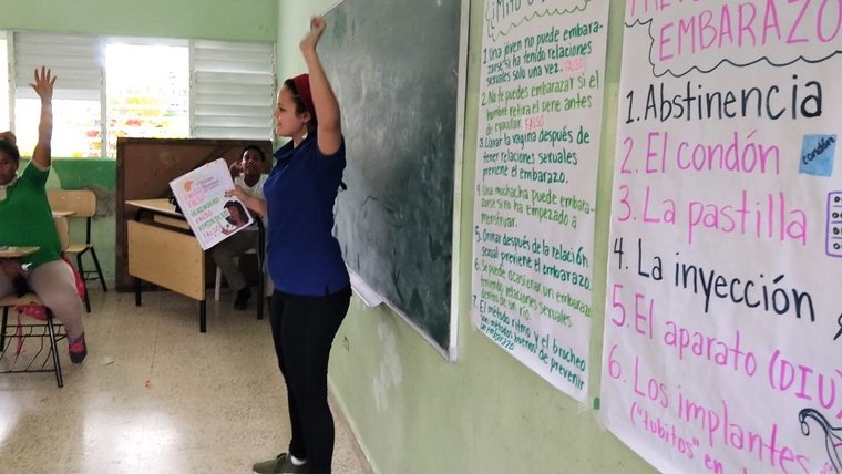 Volunteer Ashley stands in the front of a classroom and raises her hand to encourage student participation.