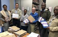 Five people, comprised of South Africans and an American, stand around a table holding paper files.