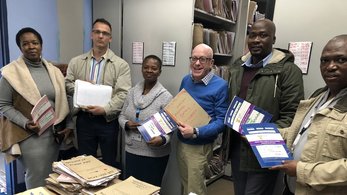 Five people, comprised of South Africans and an American, stand around a table holding paper files.