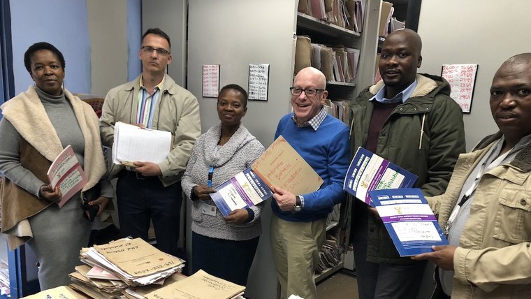Five people, comprised of South Africans and an American, stand around a table holding paper files.