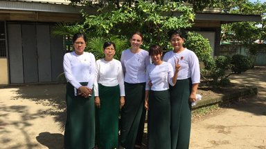 Five women teachers dressed in long green skirts and white shirts, standing outside of the school.