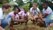 Two Peace Corps Volunteers and three Jamaican teens crouch down in a semi-circle as they plant a tree during Camp GLOW.