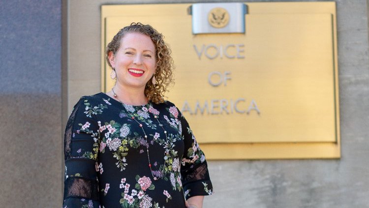 A woman leans against a hand rail and smiles. Behind her is a plaque that says "Voice of America"