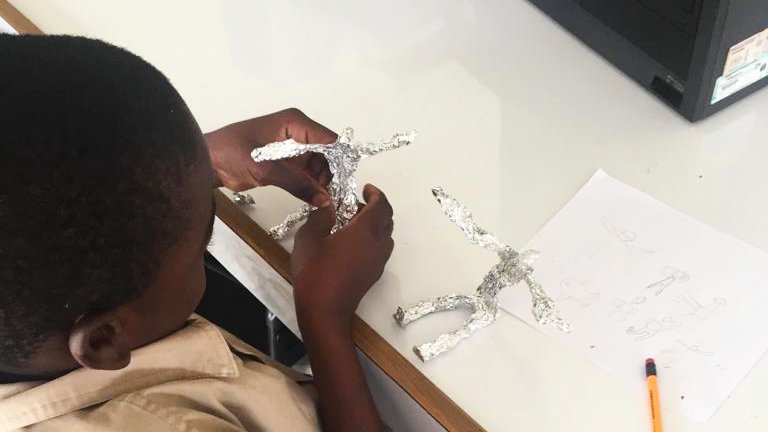 Jamaican Grade School Student sitting down and working on a sculpture of a man made out of foil in a white classroom