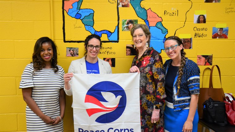 Four women stand together in a classroom holding a Peace Corps flag.