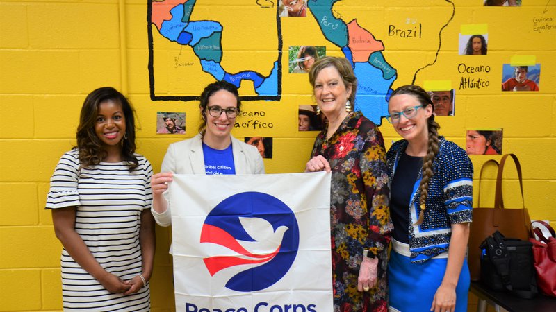 Four women stand together in a classroom holding a Peace Corps flag.