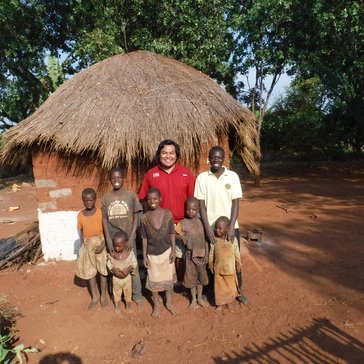 A Volunteer of Native American heritage stands in front of a Zambian home with a large family, smiling.