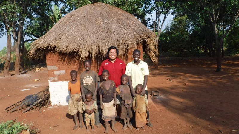 A Volunteer of Native American heritage stands in front of a Zambian home with a large family, smiling.