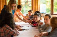 A group of young girls working on an art project at a table, while the light shines in through nearby window.