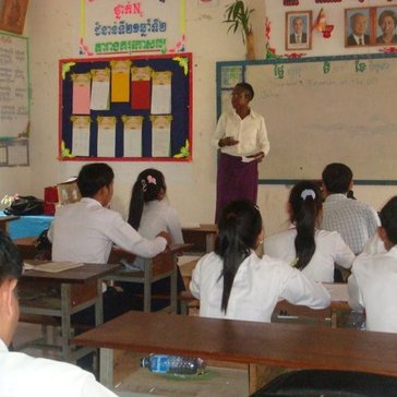 An older, Black woman stands in front of a classroom teaching Cambodian students.