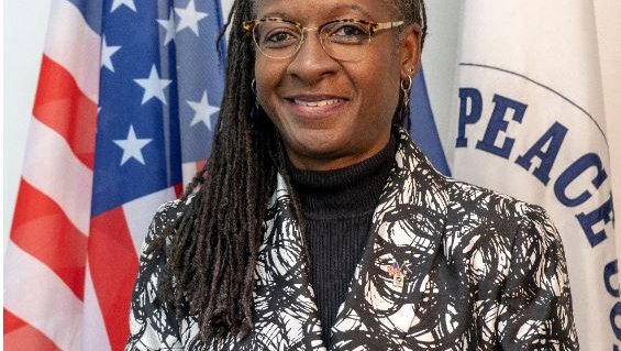 A Black woman smiles in front of an American flag and a Peace Corps flag.