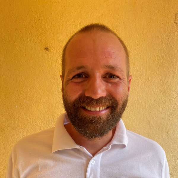 Headshot of a Volunteer smiling wearing a white shirt and standing against a yellow wall.
