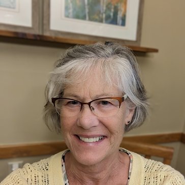 A woman in a yellow shirt and glasses with gray hair smiles.
