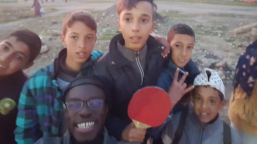 A black, male Volunteer smiles for a selfie outside with many Moroccan children